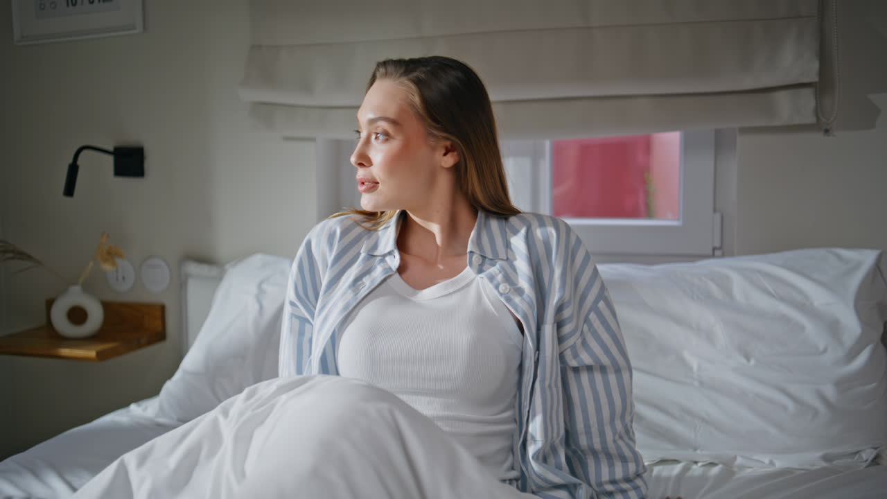 Energized woman wake up on white sheets lit by morning sunlight. Lady in pajamas