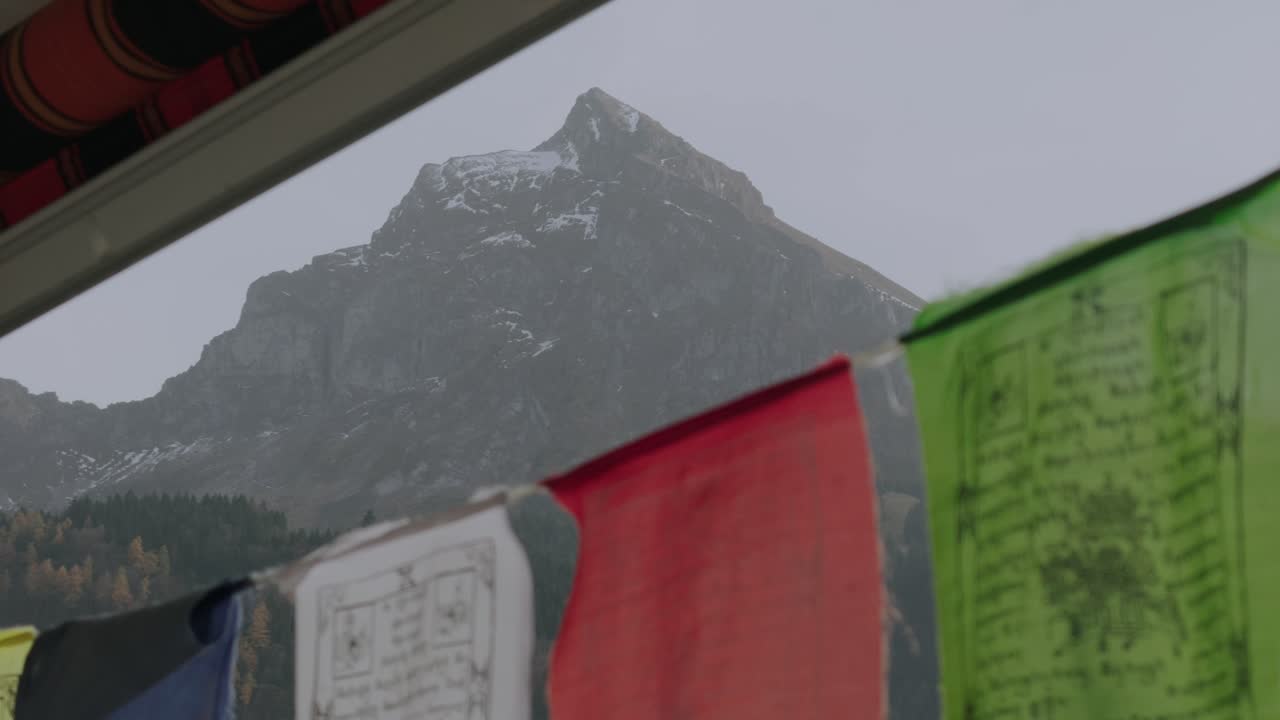 Colorful Tibetan prayer flags flutter in the wind with the Fronalpstock mountain peak in the background. Captured outdoors on a cloudy day in the Swiss Alps, creating a peaceful atmosphere