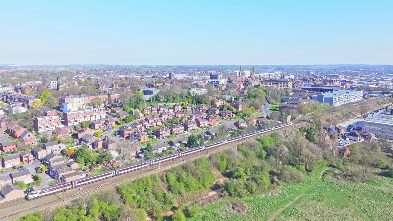 Aerial view of Wakefield, England, featuring traditional British brick homes, lush green spaces, industrial zones, and a high-speed train cutting through the suburban landscape.
