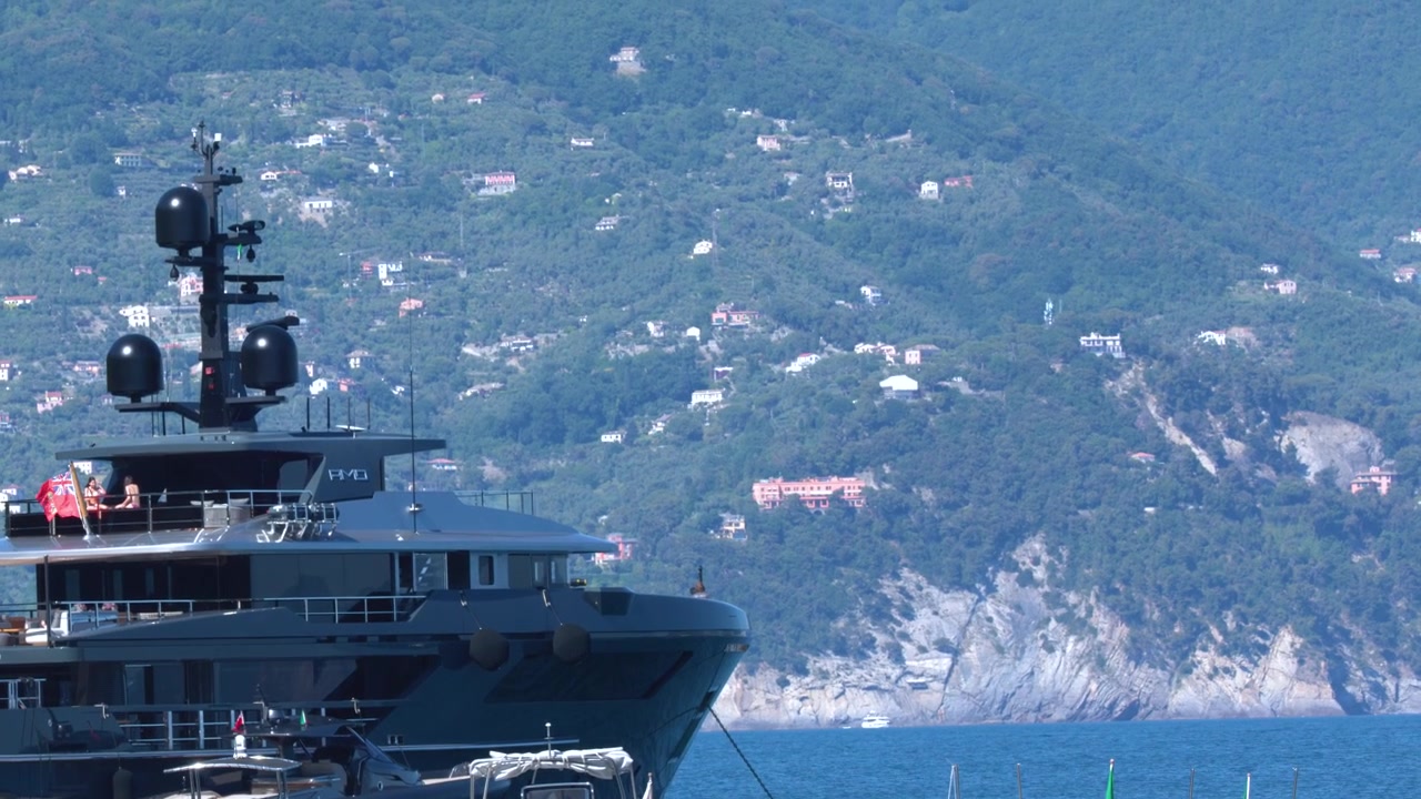 Un grande yacht di lusso scuro si trova in un porto mediterraneo sullo sfondo di rigogliose colline verdi.