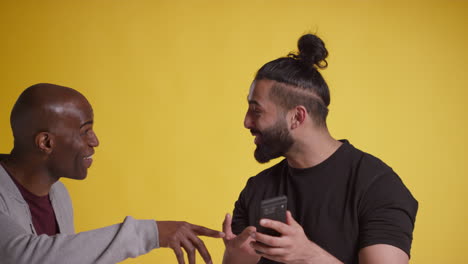 studio shot of two excited male friends with mobile phone celebrating winning money against yellow background 2