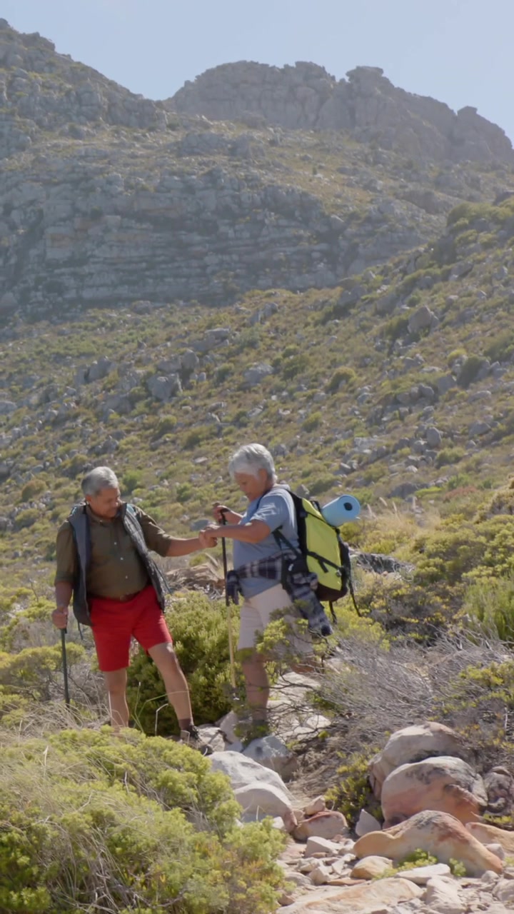 Vertical video: Senior couple descending rocky slope, stepping on loose rock, using pole to steady