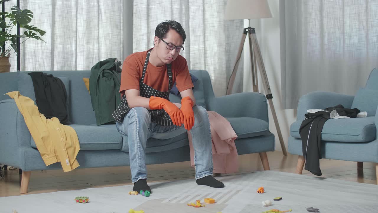 소파 에 앉아 있고, 망진창 한 방 을 청소 하는 데 피곤 하기 때문 에 두통 을 고 있는 아시안 남성 집주인