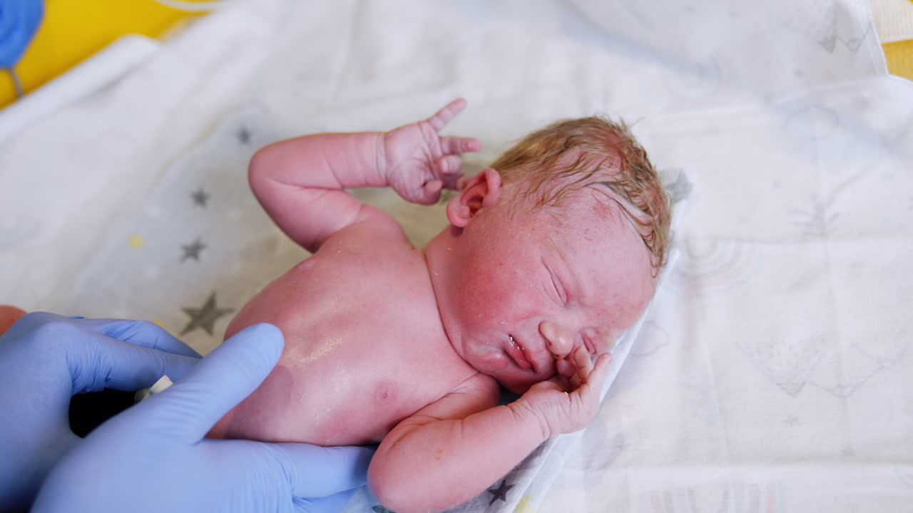 Caucasian newborn is unhappy to be disturbed by the doctor's check up. Hands in blue latex gloves press on baby belly. Close up.