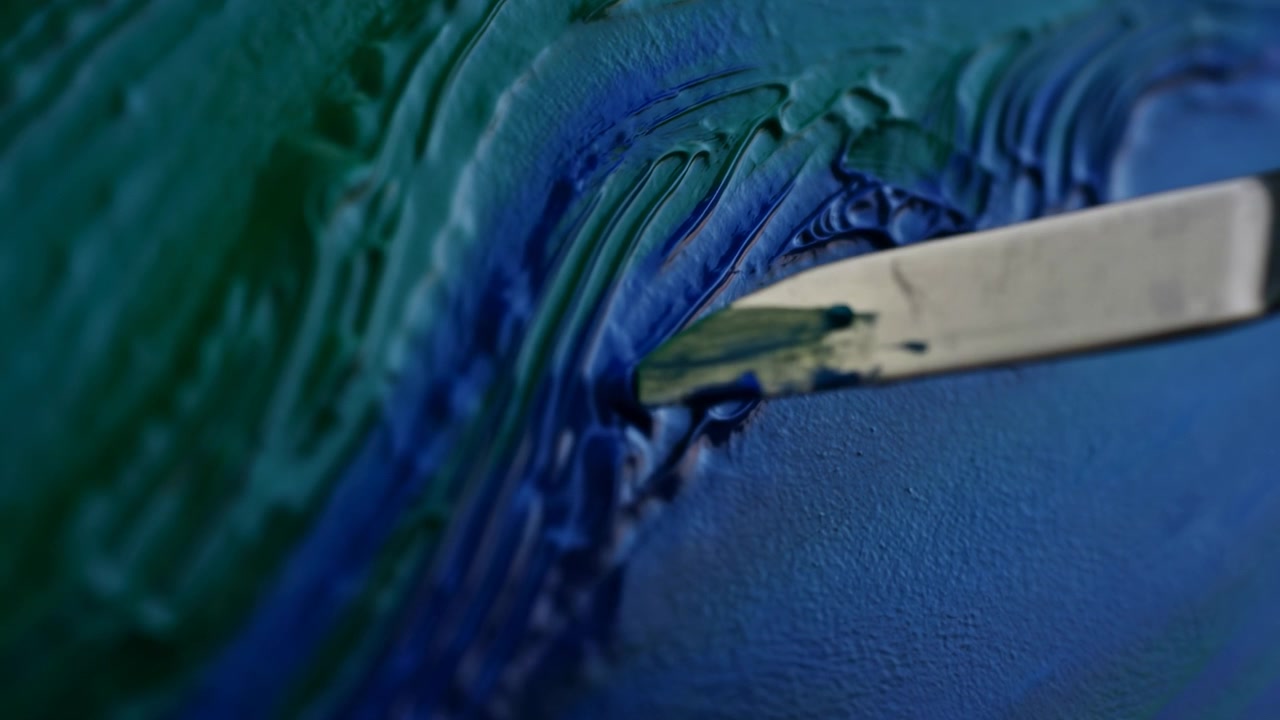 Vista ravvicinata di una spatola metallica che tocca pittura spessa blu e verde su una superficie texturizzata, trascinando il colore in creste lucide, ampie pennellate lisce, tratti curvi e dettagli astratti a strati