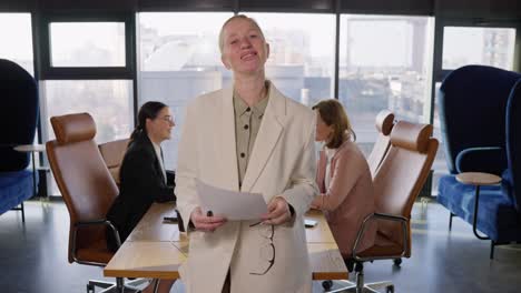 portrait of a confident middle-aged blonde woman who takes off her glasses and poses in a white business suit near her work colleagues in the office at a table in an office with large windows