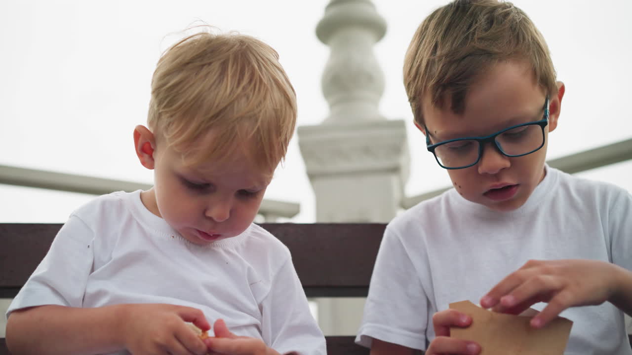 dos hermanos están sentados, profundamente centrados en los bocadillos que tienen en sus manos, sus bocas están llenas mientras disfrutan de sus golosinas
