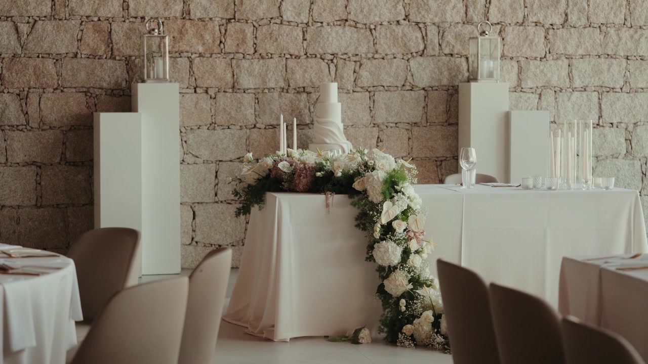White wedding cake on a draped table with a garland of white roses stands against a granite wall in a reception hall.
