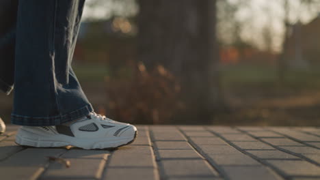 a close-up shot of a person wearing jeans and white shoes, carefully wandering through a park. the warm, soft light of the setting sun adds a reflective