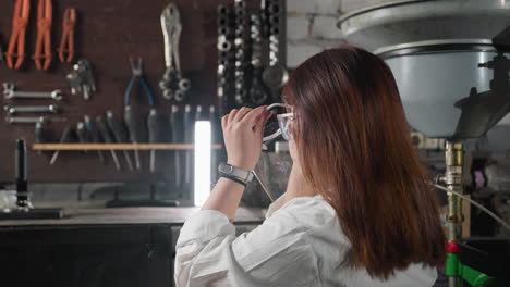 inventor in white coat picks up protective face shield, puts it on, and relaxes back on seat, with smart watch visible on left hand, surrounded by light, machinery, and equipment in workshop