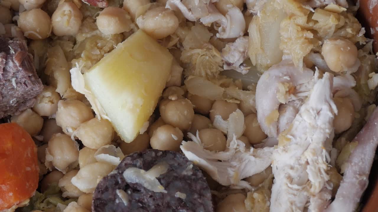 Slow pan right revealing Spanish cocido stew with chickpeas, meat, chorizo, potatoes, and vegetables in a clay dish