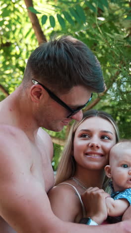 family of three on simmer vacation. topless man wearing glasses embraces his wife holding a child. vertical video