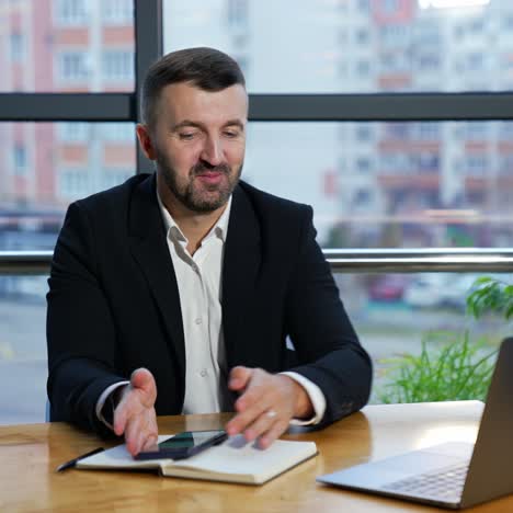 Man in a jacket and suit talk to someone on the laptop and holds a smartphone. Video call to the partners in office. Negotiations via laptop
