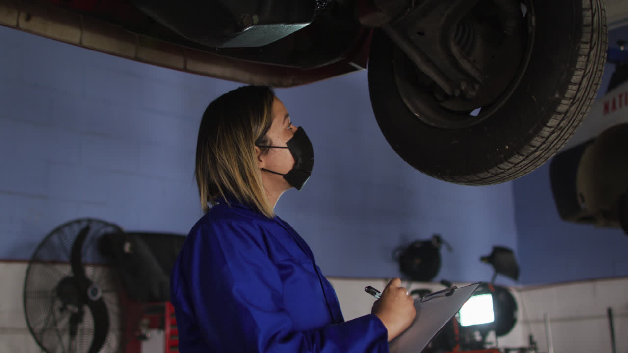 mecánico femenino con máscara facial sosteniendo el clipboard e inspeccionando el coche en una estación de servicio de coches