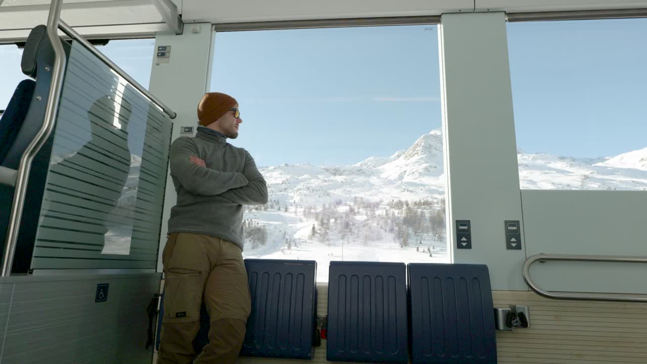 Junger Mann steht an einem Zugfenster und blickt an einem sonnigen Wintertag nach draußen auf schneebedeckte Berge.