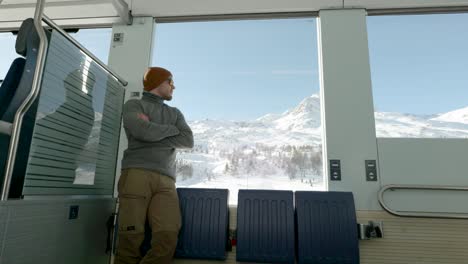 Young-caucasian-man-standing-next-to-a-train-window-and-looking-outside-to-snow-covered-mountains-during-a-sunny-winter-day