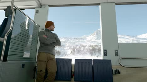 young caucasian man standing next to a train window and looking outside to snow covered mountains during a sunny winter day