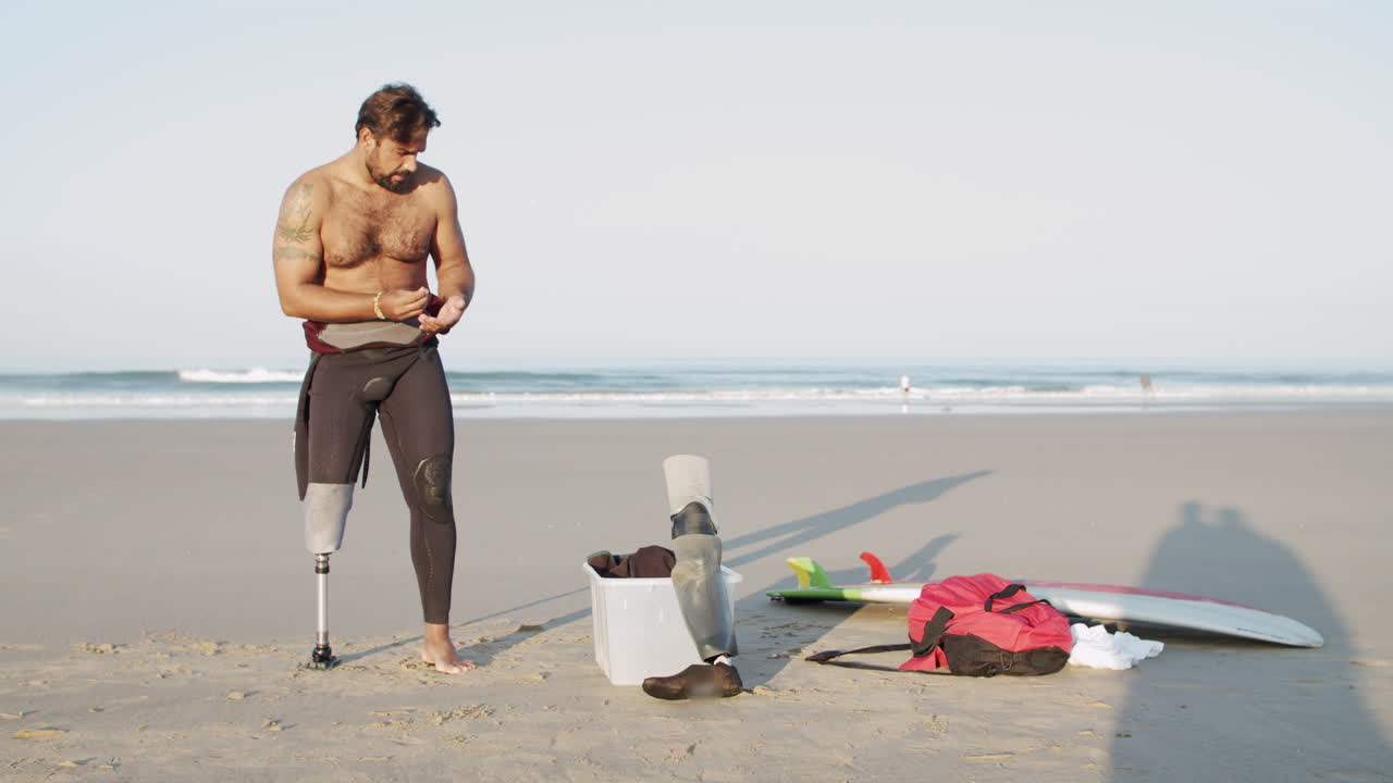 front view of bearded disabled surfer with naked torso standing on  coastline, wearing wetsuit, taking watch and bracelet off and preparing for  training