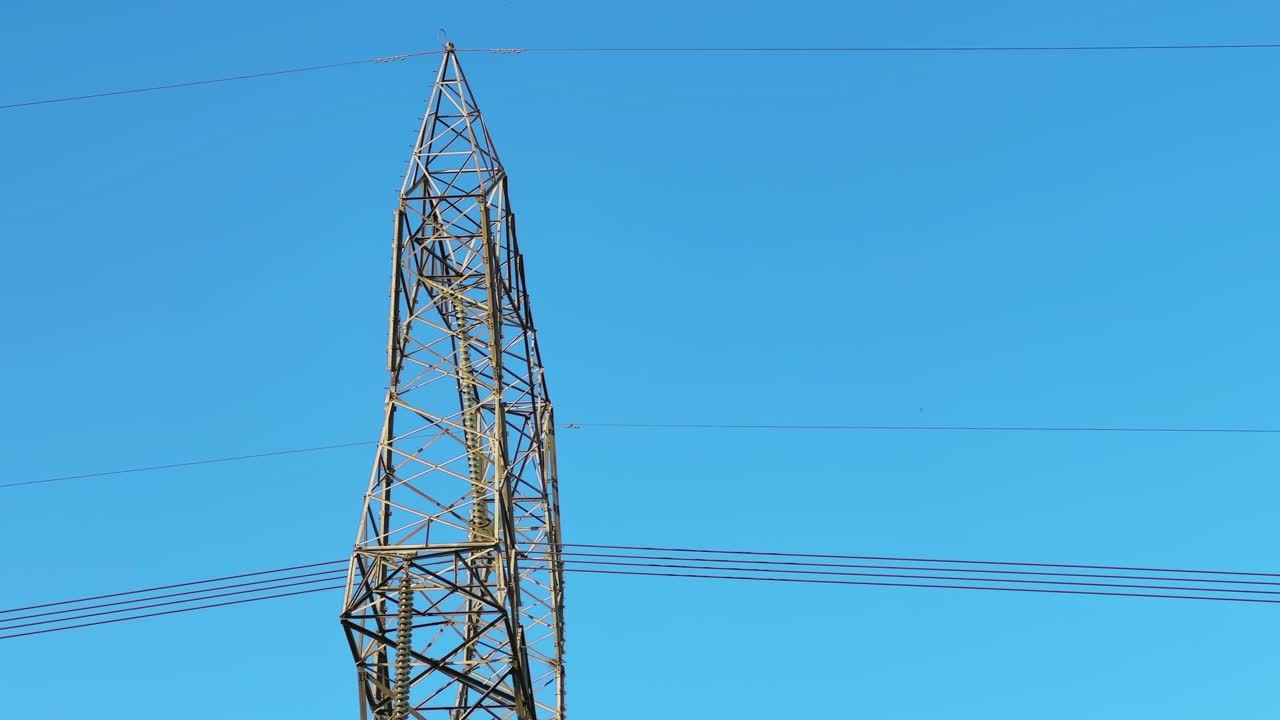 Close side view of a steel transmission tower against clear blue sky showing lattice structure and power lines highlighting energy infrastructure scale and engineering