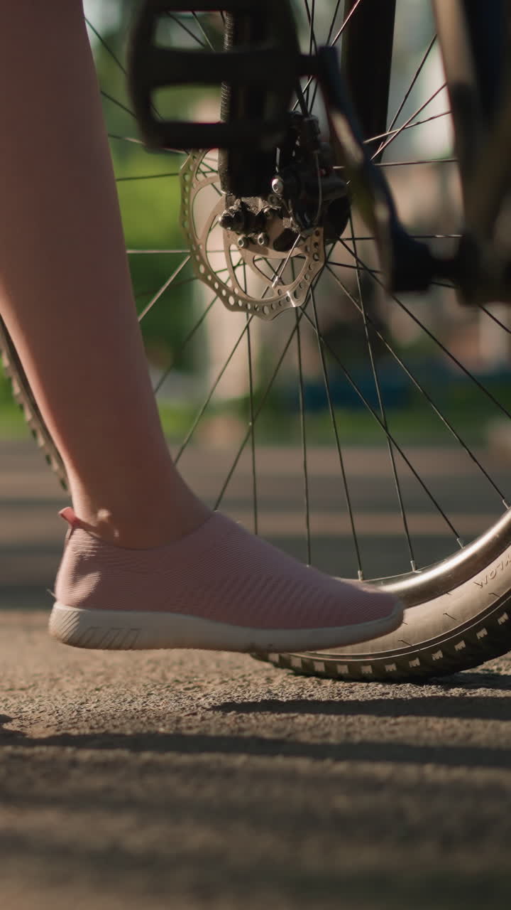 핑크색 스니커를 특징으로 한 자전거 타이어의 개별적인 공기 압력을 발로 확인하는 다리 시각, 배경은 멀리 있는 건물과 함께 울창한 초록색과 나무를 보여줍니다.