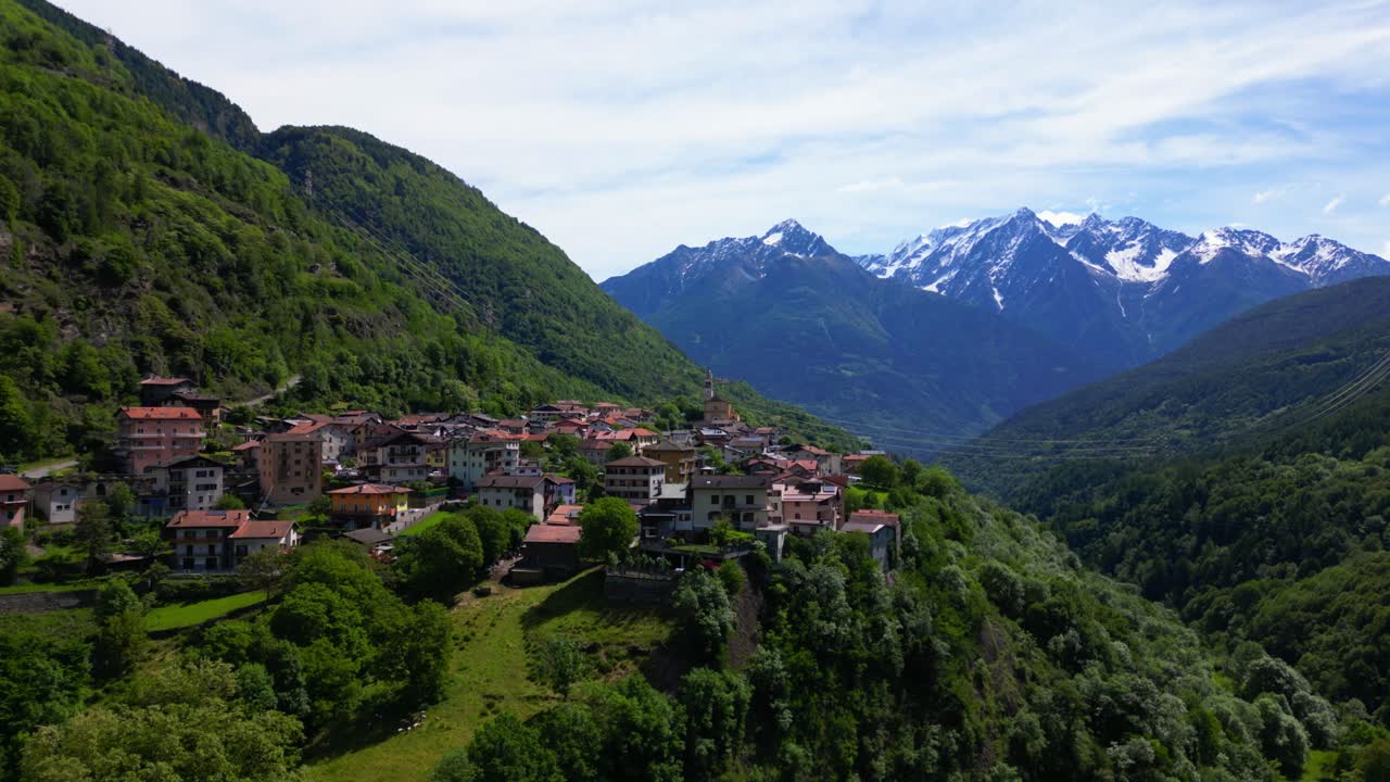 Drone view of terracotta-roofed homes clustered on a lush slope with dramatic snow-covered peaks behind. Shot at Malonno, Italy (Italia)