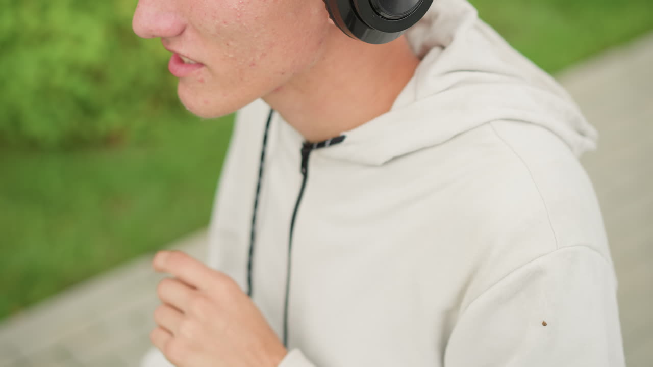 흐린 날씨의 공원 산책로에서 헤드폰을 테스트하며, 깔끔한 언더컷과 후드티를 입고, 집중한 표정으로 헤드밴드를 착용하고 착용감을 조절하며 사운드를 확인하면서 길을 따라 걷는 젊은 백인 남성