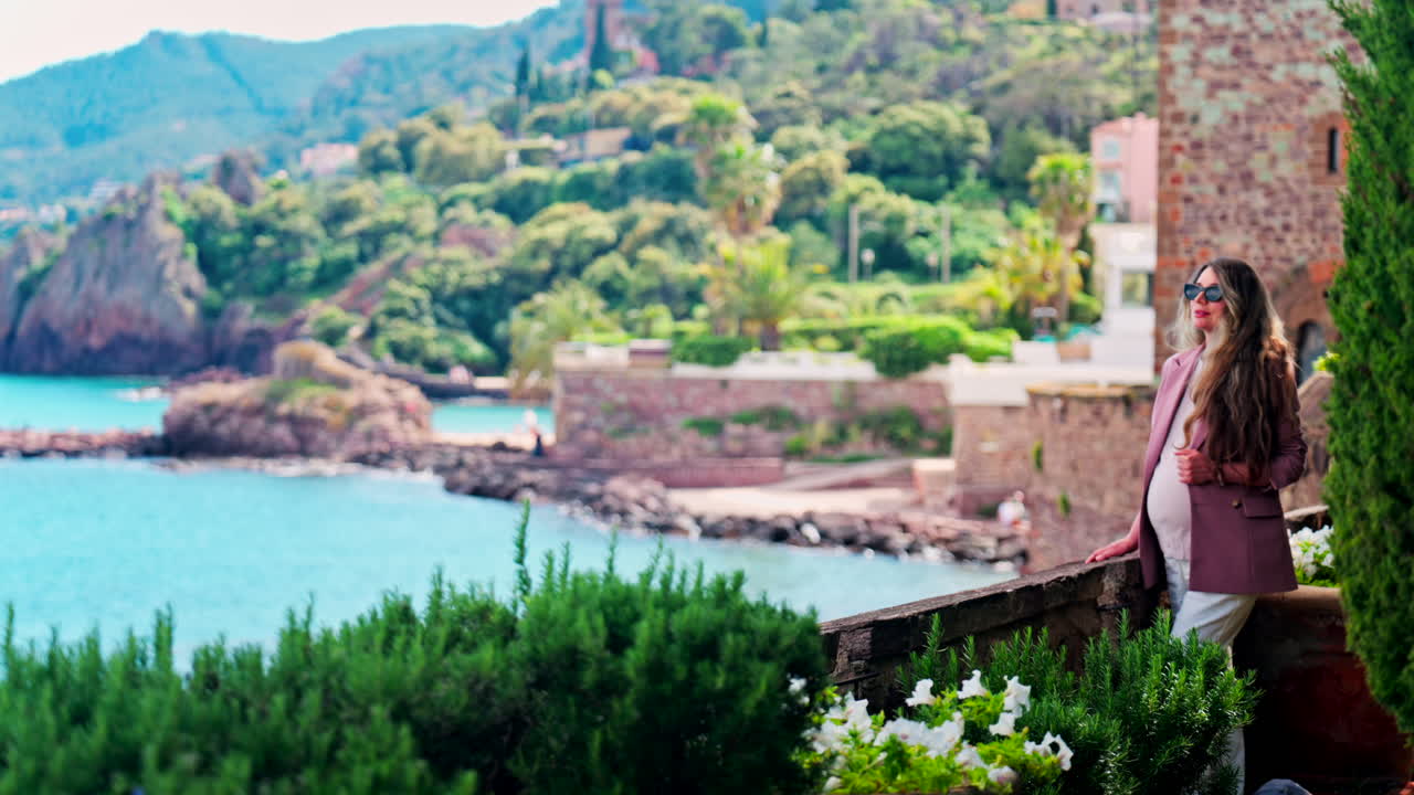 Pregnant woman with long hair, wearing sunglasses standing with the Chateau de la Napoule Castle in Mandelieu-La Napoule, France in the background