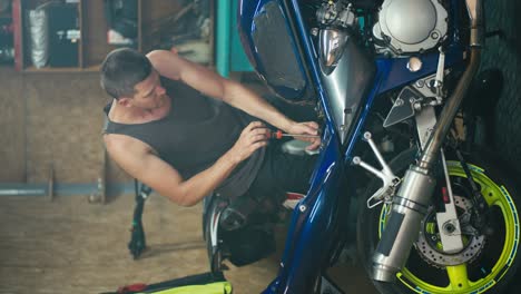 Vertical-video-of-a-male-master-Motor-mechanic-with-a-short-haircut-in-a-gray-T-shirt-repairing-his-blue-and-yellow-bike-and-dismantling-it-into-pieces-in-his-workshop-garage