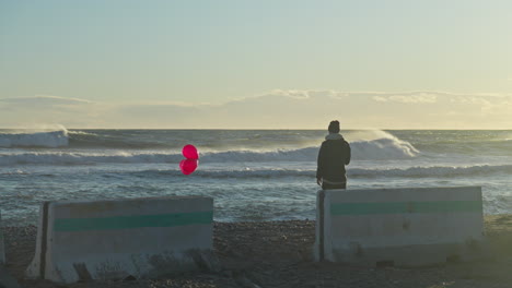 a man holds three red balloons as he watches the ocean waves break before him