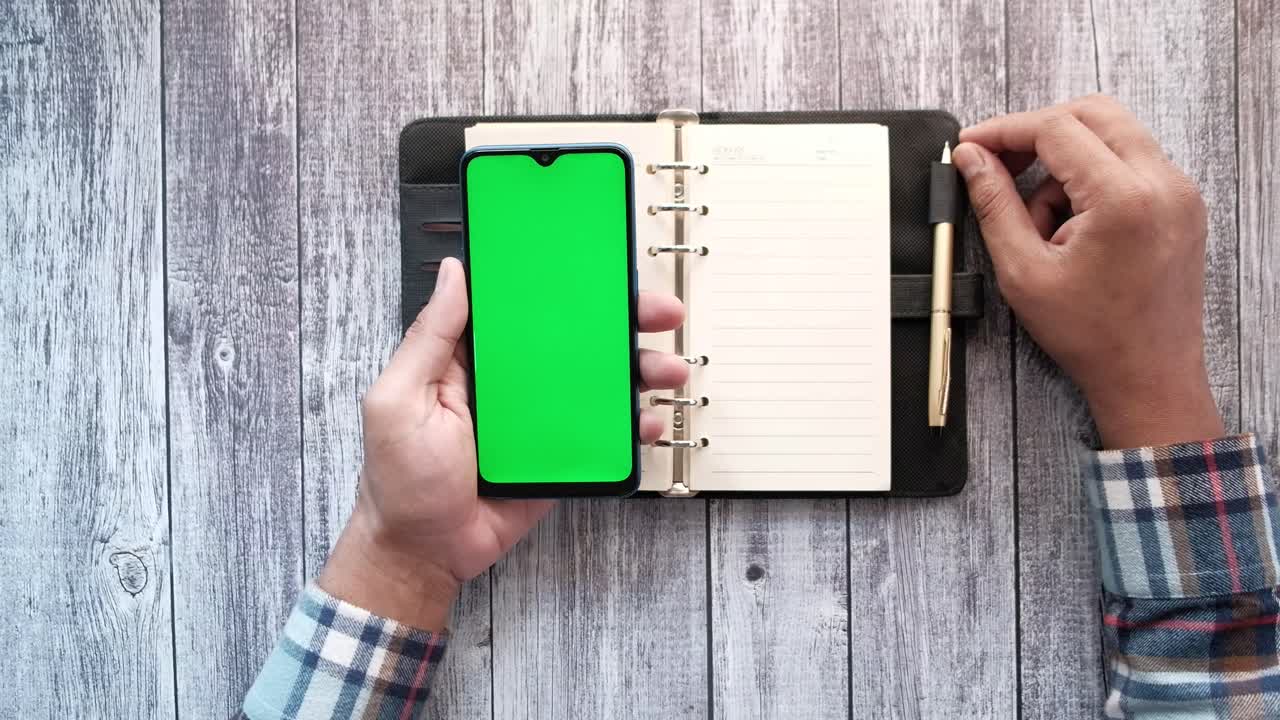 Person holding a smartphone over a notebook and pen on wooden table