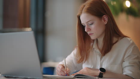 Una-Joven-Estudiante-Está-Haciendo-Su-Tarea-Escribiendo-En-Un-Cuaderno-Buscando-Información-En-Internet.