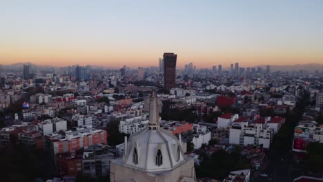 Amplias-Tomas-De-Drones-Del-Enorme-Estatus-De-Jesús-Que-Se-Eleva-Sobre-La-Ciudad-De-México-Al-Atardecer,-Con-La-Parroquia-Del-Purismo-Corazon-De-Maria-Y-Edificios-En-La-Parte-De-Atrás