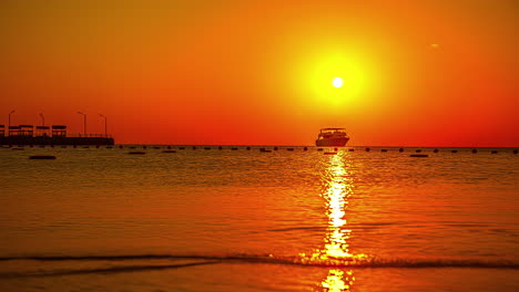 Zeitrafferaufnahme-Des-Orangefarbenen-Himmels-Bei-Sonnenuntergang-Mit-Einem-Schiff-Auf-Dem-Meer-Und-Wandernden-Menschen-Auf-Der-Promenade