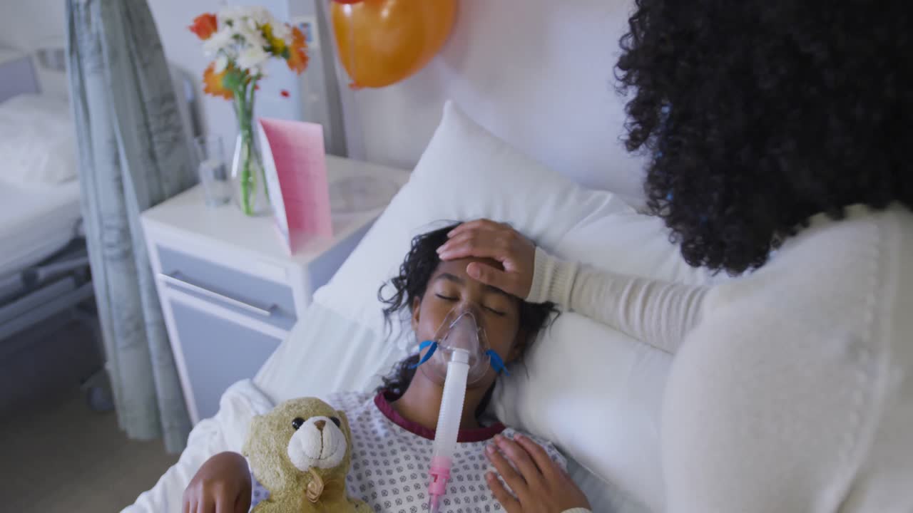 Madre afroamericana che si prende cura della figlia con maschera per l'ossigeno sdraiata a letto in ospedale.