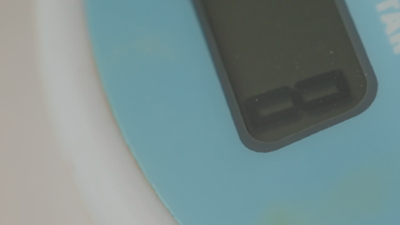 Display ravvicinato di una bilancia digitale che mostra cifre che cambiano su un pannello blu, messa a fuoco macro sui segmenti LCD e superficie ruvida, illuminazione soffusa e diffusa, ispezione ravvicinata della lettura, chef che misura gli ingredienti