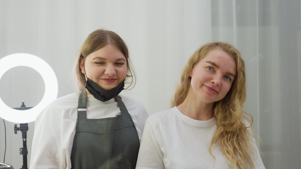 Client and beautician stand in salon smiling warmly at camera, technician gently pulling down black mask in relaxed gesture, surrounded by beauty tools and glowing ring light on white vanity