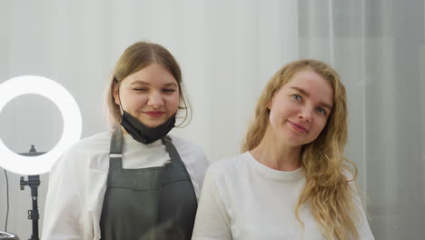 client and beautician stand in salon smiling warmly at camera, technician gently pulling down black mask in relaxed gesture, surrounded by beauty tools and glowing ring light on white vanity