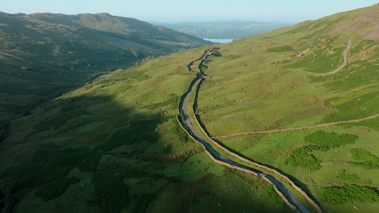 Mountain pass road lined by stone wall in early morning sunshine with lake Windermere in distance. Summer. Kirkstone Pass, Lake District, Cumbria, UK