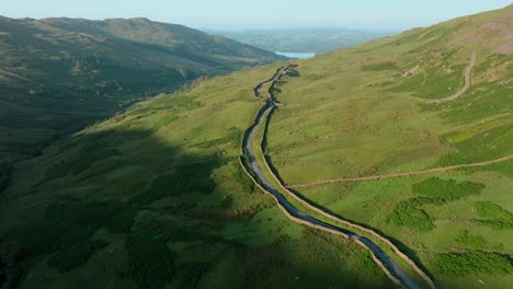 mountain pass road lined by stone wall in early morning sunshine with lake windermere in distance. summer. kirkstone pass, lake district, cumbria, uk