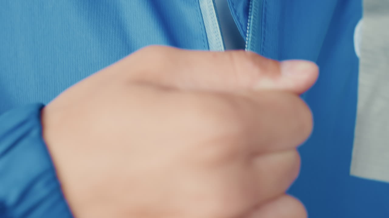 Close up hands zipping blue jacket, football player preparing for rainy match, tight framing on zipper sleeve and thumb movements, water droplets on fabric, determined pregame focus