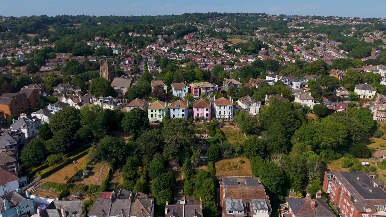 Inquadratura di apertura con drone in ascesa che si eleva oltre una fila di case a schiera color pastello su una collina a Hastings, rivelando la città più ampia e i tetti che si estendono in lontananza oltre gli alberi.
