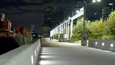 Muelles-De-Brooklyn-Por-La-Noche-Con-El-Horizonte-De-Nueva-York-Al-Fondo,-Junto-A-Un-Parque-Con-Grandes-Matorrales-Verdes,-En-Un-Día-Ventoso,-Con-Un-Gran-Barco-Industrial-Amarrado-Junto-Al-Muelle-De-Cemento