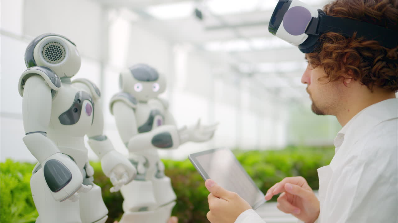 Laboratory technician in a white coat wearing virtual reality headset analysing graphs on a tablet while interacting with two humanoid robots in a greenhouse farm