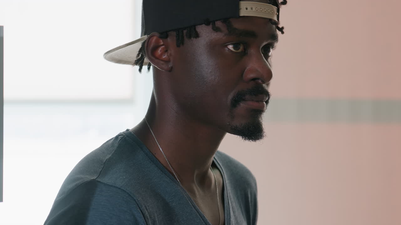 Side view of young man in casual clothing and backward cap resting near metal pole, gazing forward with calm focused expression in softly lit indoor space