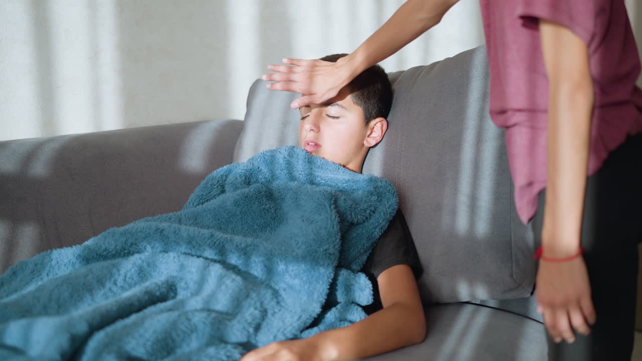 Little boy lying on couch with fever, wrapped in blanket, being cared for, temperature checked by someone nearby, with soft sunlight filtering through window, casting shadows