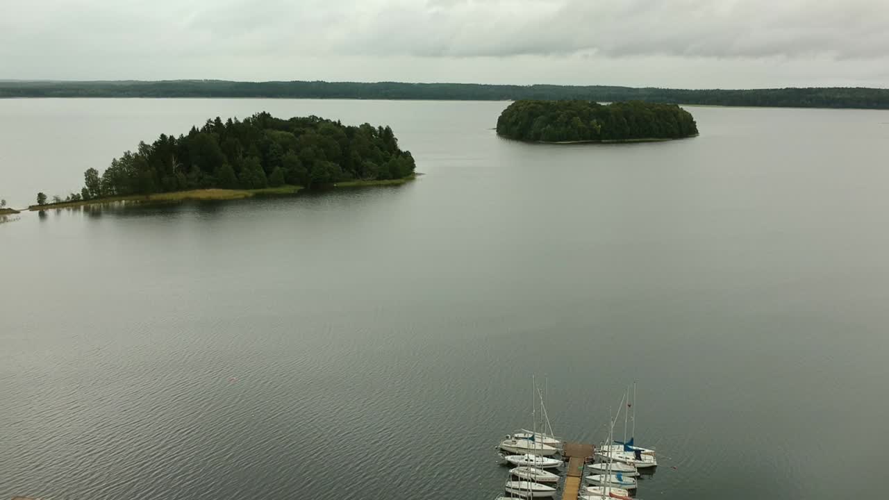Ujęcie z lotu ptaka cofające się od małych wysp na jeziorze Plateliai na Litwie, ukazujące pomost, budynek i zaorane pole.