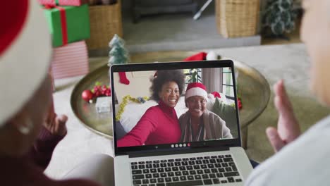 diversas amigas mayores agitando y usando una computadora portátil para una videollamada de navidad con la familia en la pantalla