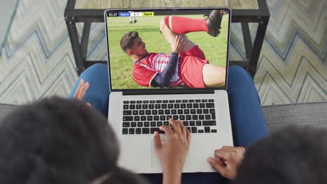 Video-of-african-american-couple-sitting-on-sofa-and-watching-football-on-laptop-at-home