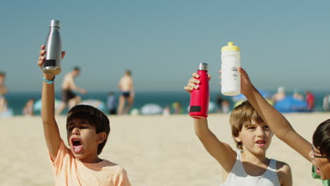 Happy-sportsmen-sitting-on-beach-and-raising-bottles-of-water-up