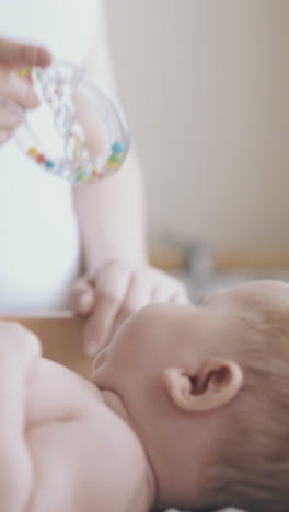 adorable infant son in white diaper lies on changing table and enjoys leg massage and toy in mother hand close view